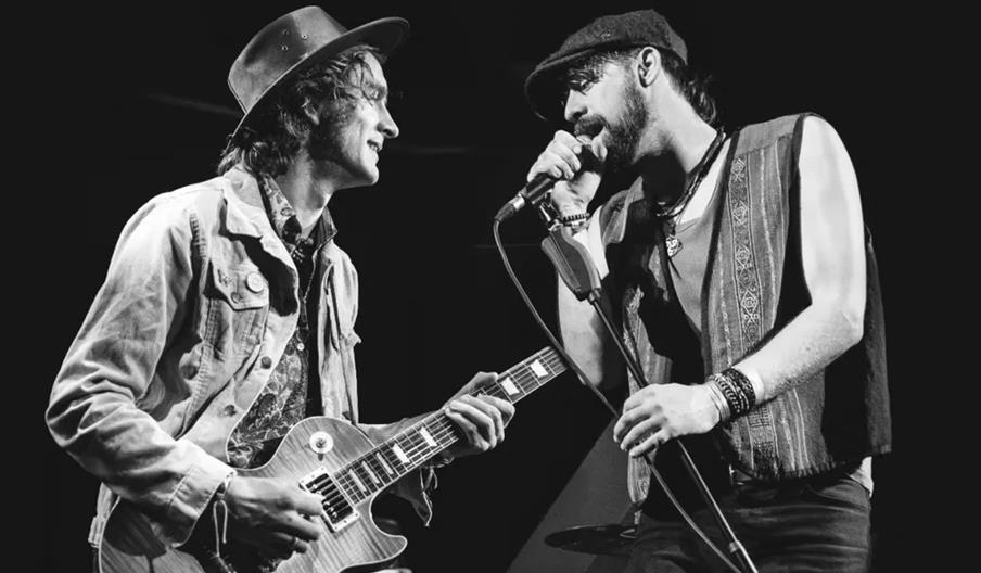 Black and white image of two musicians performing on stage; one is playing an electric guitar and wearing a hat and jacket, while the other is singing