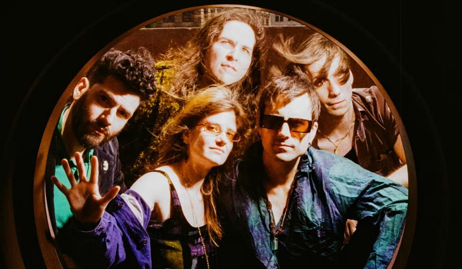 Five people pose closely together, looking through a round window or porthole, with varied expressions and casual, retro-style clothing. The lighting