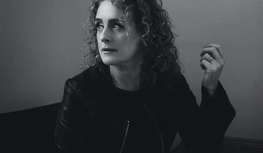 Black and white photo of a woman with curly hair sitting at a table, looking thoughtfully to the side, with a cup of coffee in front of her.