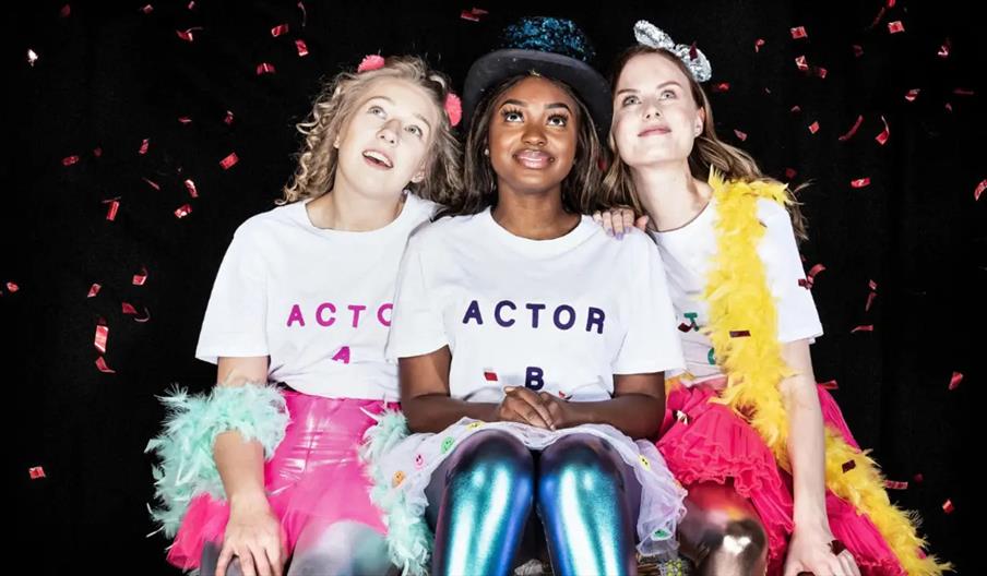Three young women in colorful costumes sit closely together on stage, wearing white shirts labeled "ACTOR A," "ACTOR B," and "ACTOR C" and looking upw