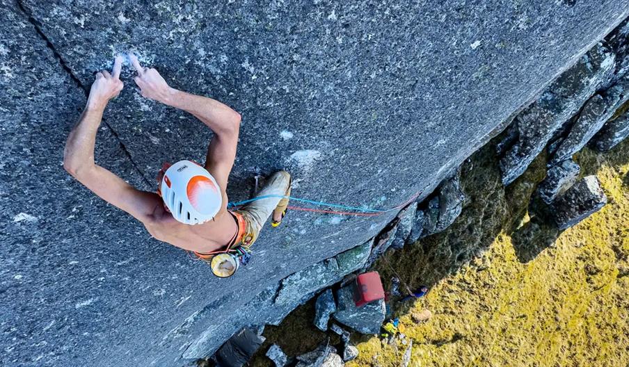 A climber wearing a helmet and harness grips a vertical rock face, viewed from above, with ropes attached for safety and grassy ground far below.