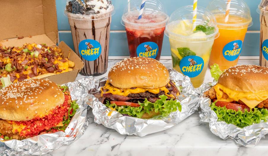 3 cheeseburgers in foil, a chocolate shake, strawberry drink, lemon drink and orange drink in plastic cups with straws all on a marble table.