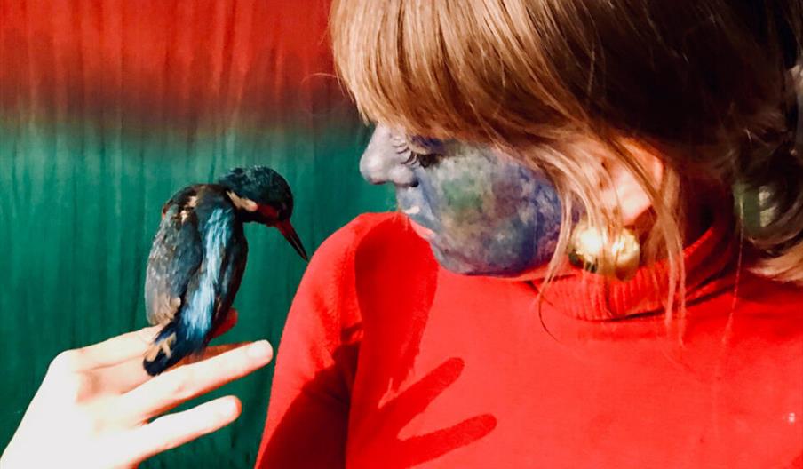 A woman in a bright red turtleneck with blue paint on her face looks at a small blue bird perched on her fingers, against a red and green backdrop.