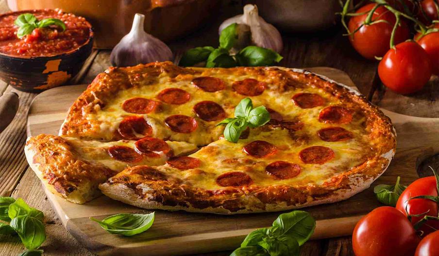 A pepperoni pizza on a wooden paddle, with fresh garlic, herbs, tomatoes and tomato sauce around it,