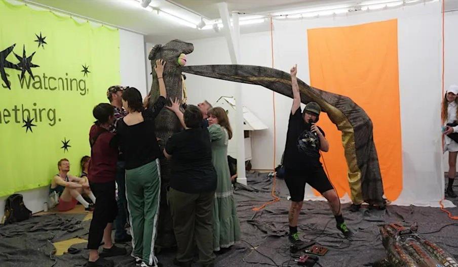 A group of people interact with a large, sculptural puppet resembling a dinosaur or dragon in an art gallery. One person stands in front with a microp