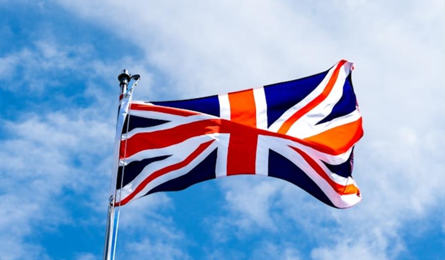 The Union Jack flag of the United Kingdom is flying on a flagpole against a bright blue sky with scattered clouds.