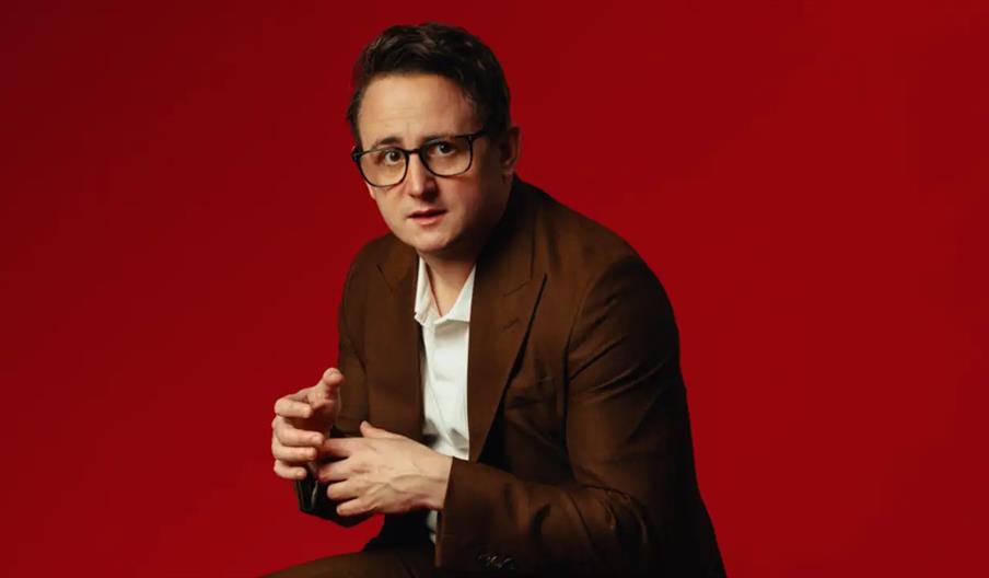 A person with short dark hair and glasses, dressed in a brown suit and white shirt, sits against a solid red background, looking slightly to the side