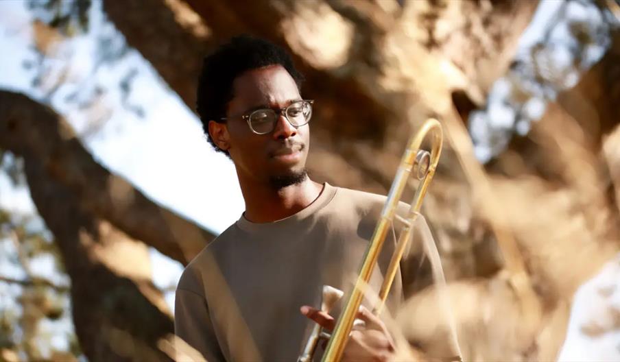 A young man wearing glasses and a beige shirt stands outdoors, holding a trombone, with trees and sunlight in the background. Dry grass is visible in