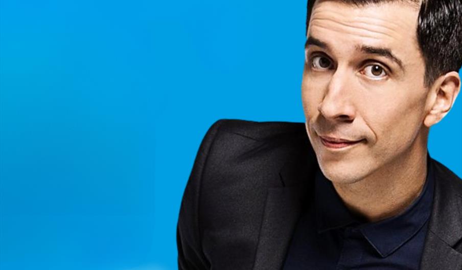 Russel Kane is wearing a navy shirt and black blazer is looking at the camera with a slight smile, set against a bright blue background with clouds in