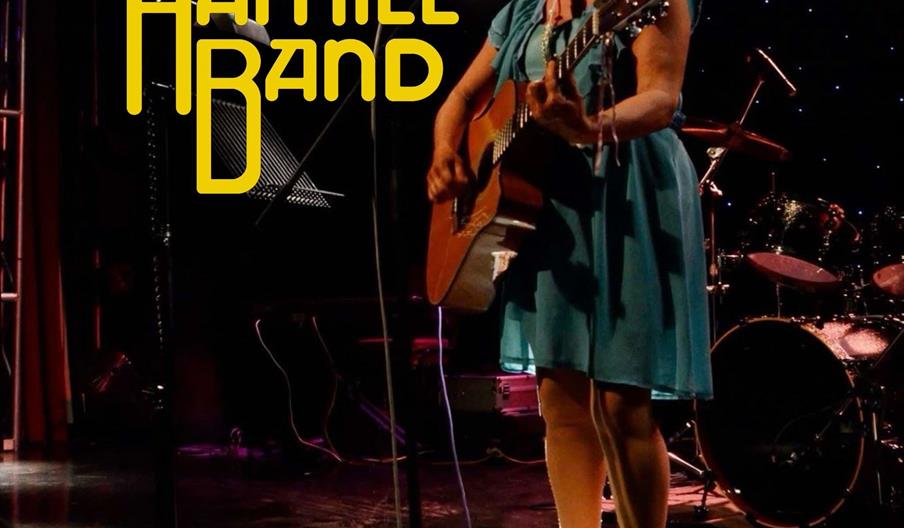 A woman in a teal dress plays an acoustic guitar and sings into a microphone on stage under spotlights, with a drum set in the background. Bold yellow