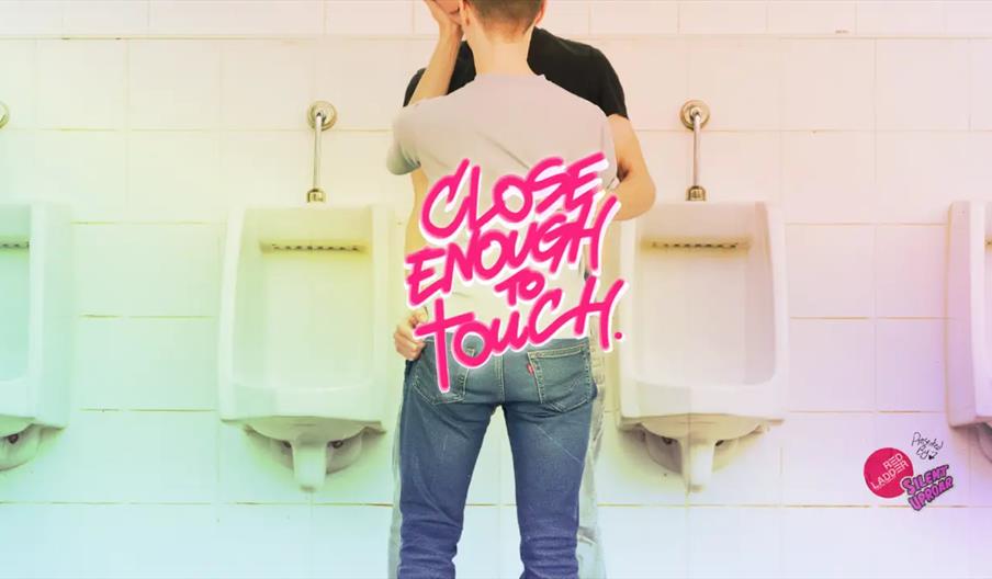 Two men stand close together and kiss in front of urinals in a public restroom. Overlaid text in bright, bold letters reads "Close Enough to Touch."