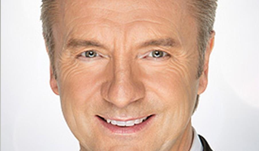 Christopher Dean smiling with short, neatly styled light brown hair wears a black tuxedo, white shirt, and black bow tie against a light, neutral back