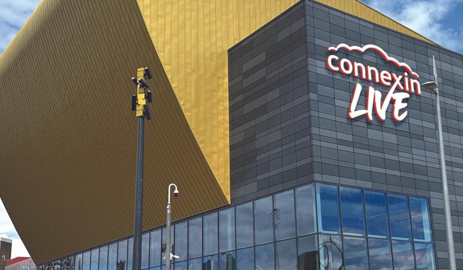 The exterior of Connexin Live in Hull, a large building with black brickword, large glass panels and a large yellow roof. Connexin Live appears on the