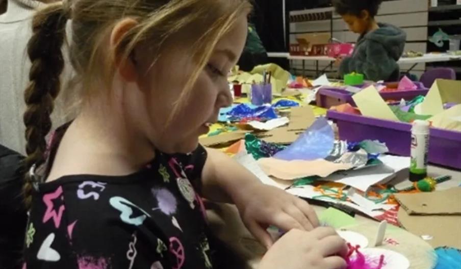 A young girl with a braid focuses on a craft project, using colorful materials at a table filled with paper, glue, and art supplies. Another child is