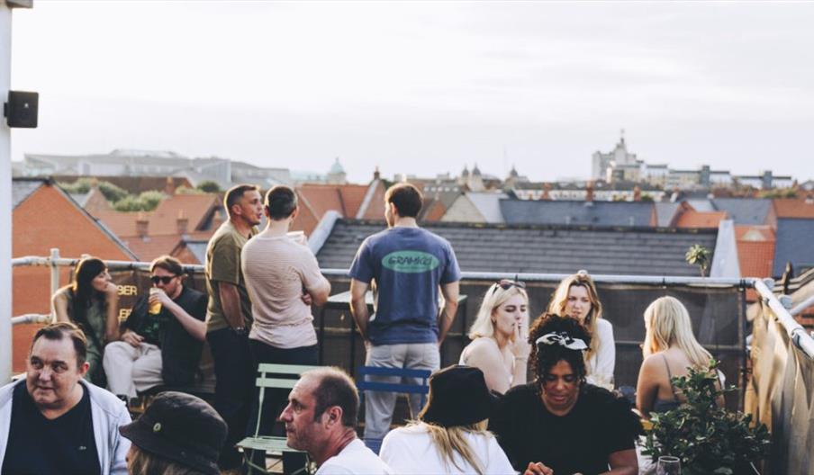 A group of people socialize on a rooftop terrace at Humber Street Gallery, with colorful rooftops and a cityscape visible in the background under a br