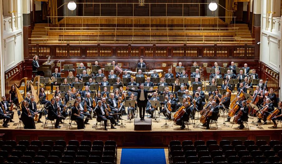 A full symphony orchestra sits on stage in a large, elegant concert hall, with musicians holding various instruments and a conductor standing at the p
