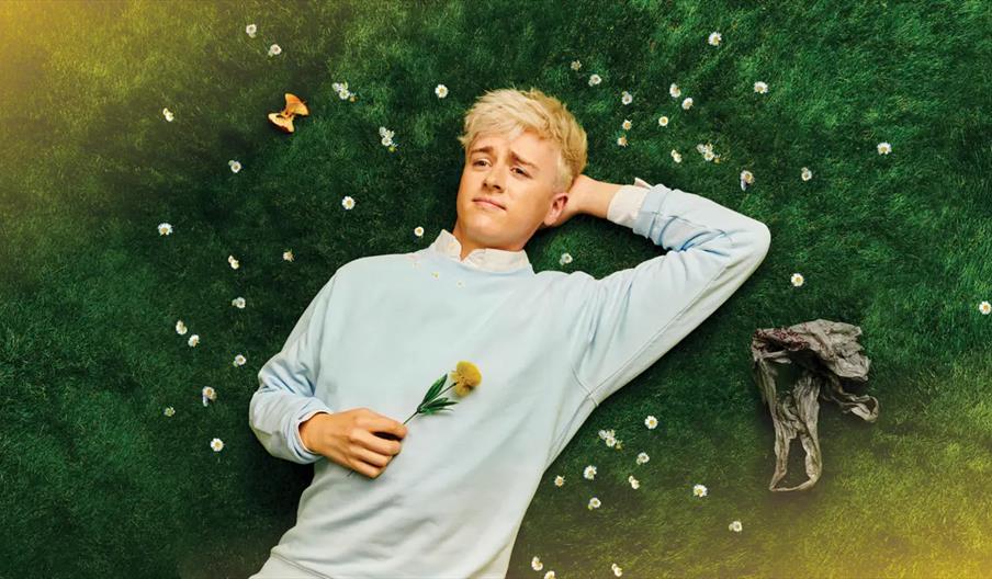 A young person with blond hair wearing a light blue sweater lies on green grass scattered with small white flowers, holding a yellow flower and gazing