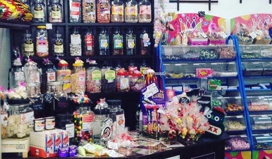 A colorful vintage-style candy shop interior filled with jars of sweets and treats on dark wooden shelves behind a counter, with additional bins of ca
