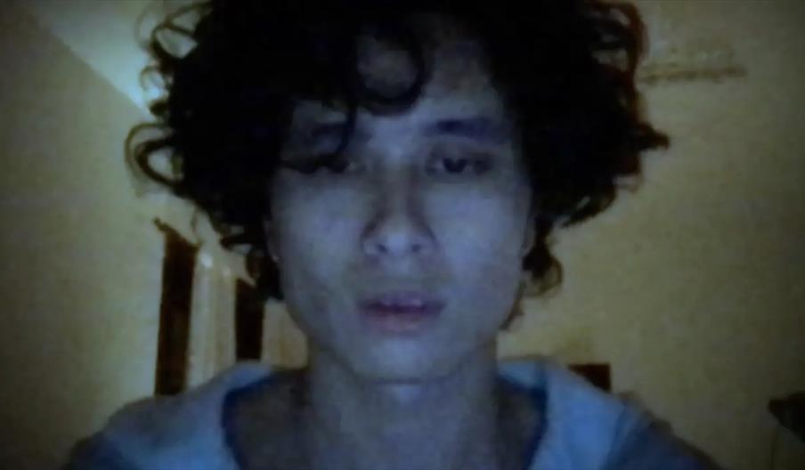 A person with curly dark hair is shown in a dimly lit room, looking directly at the camera with a neutral expression. The lighting casts a bluish tint