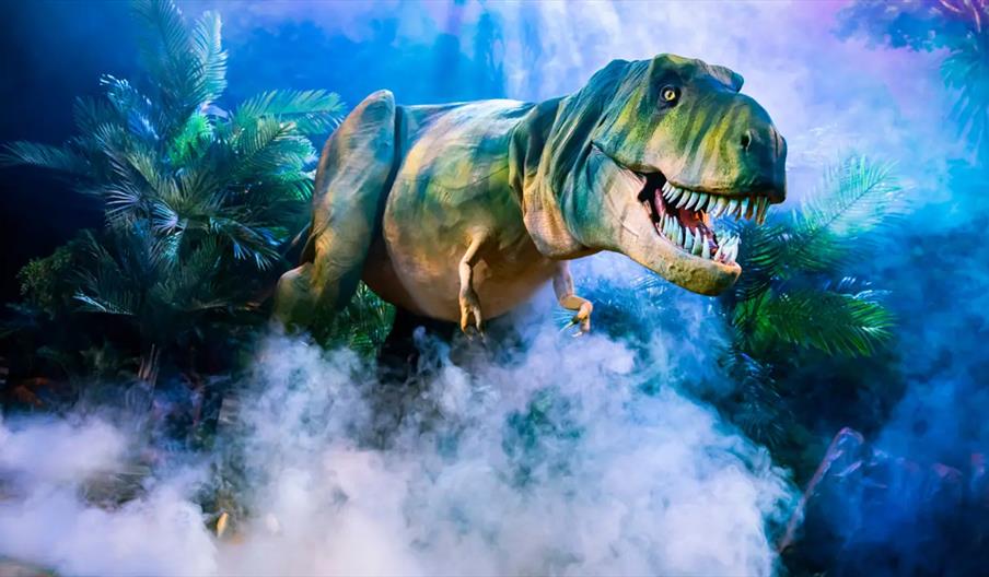 A realistic model of a Tyrannosaurus rex stands in a misty, jungle-like setting with ferns and palm leaves, its mouth open to reveal sharp teeth. Blue