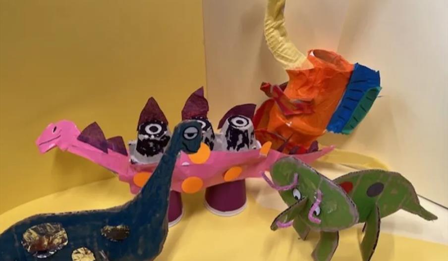 Four colourful dinosaur sculptures made from painted cardboard, egg cartons, and craft materials are displayed on a yellow background. Each dinosaur i