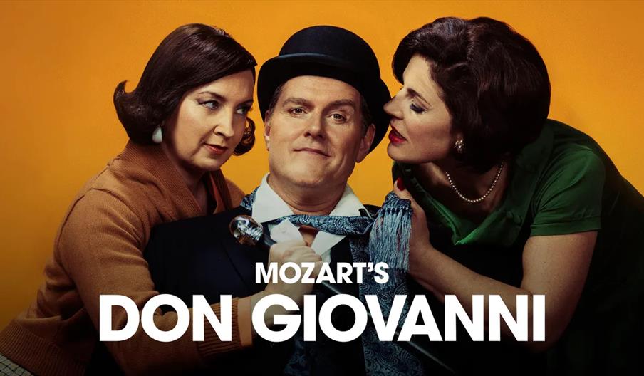Three people pose against an orange background: a man in a hat and suit holding a cane sits in the center, with two women leaning closely toward him o