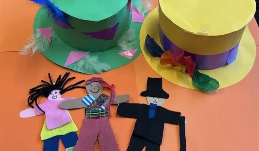 Two colorful paper hats—one green with feathers and pink triangles, the other yellow with a purple band and tissue paper flowers—sit on an orange back