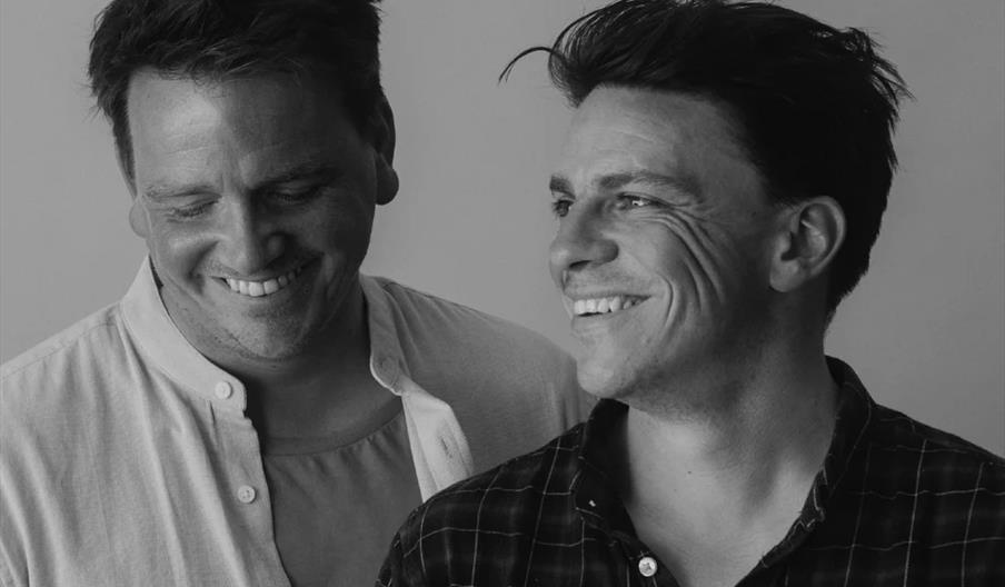 Black and white photo of two men standing side by side, both smiling and looking at each other, with one wearing a light shirt and the other wearing a