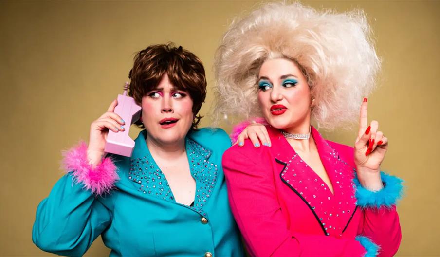Two people in colorful, retro 1980s-style outfits pose together against a plain background; one wears a teal jacket with pink fluffy cuffs and holds a