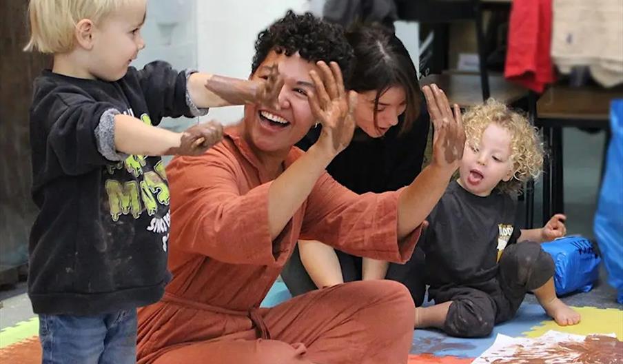 A smiling adult and three young children with paint-covered hands play together on a colorful foam mat, creating brown paint art on a long sheet of pa