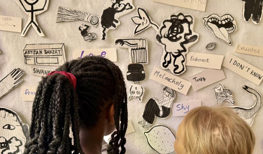 Two children stand with their backs to the camera, looking at a display of black-and-white cartoon-style drawings and handwritten words like "Melancho