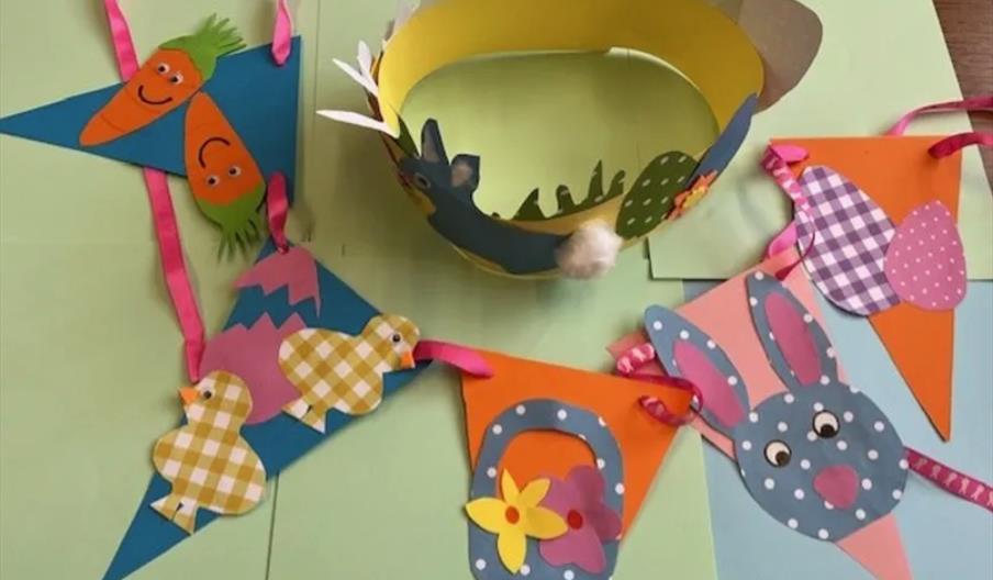 Colourful paper Easter crafts including a yellow crown decorated with cut-out shapes and a bunting made of triangular flags featuring carrots, chicks,