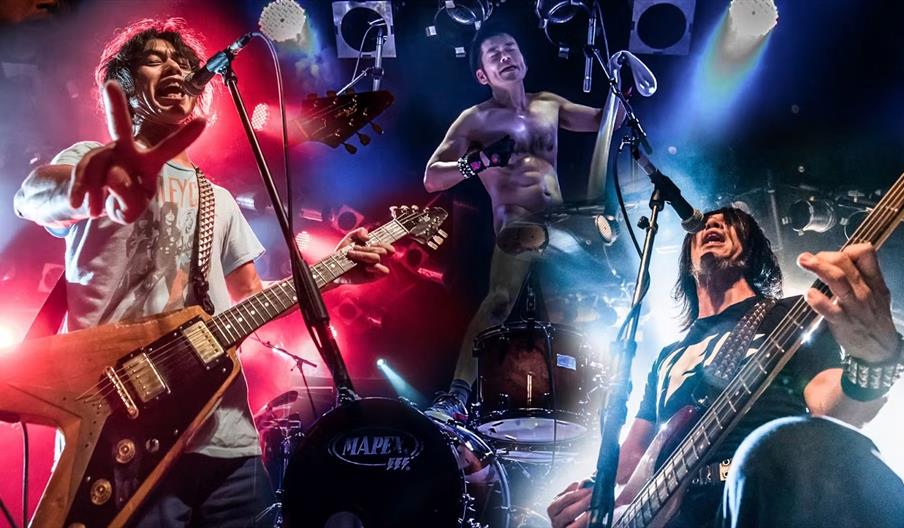 Three rock band members perform energetically on stage under dramatic colored lights; the guitarist on the left flashes a peace sign, the shirtless dr