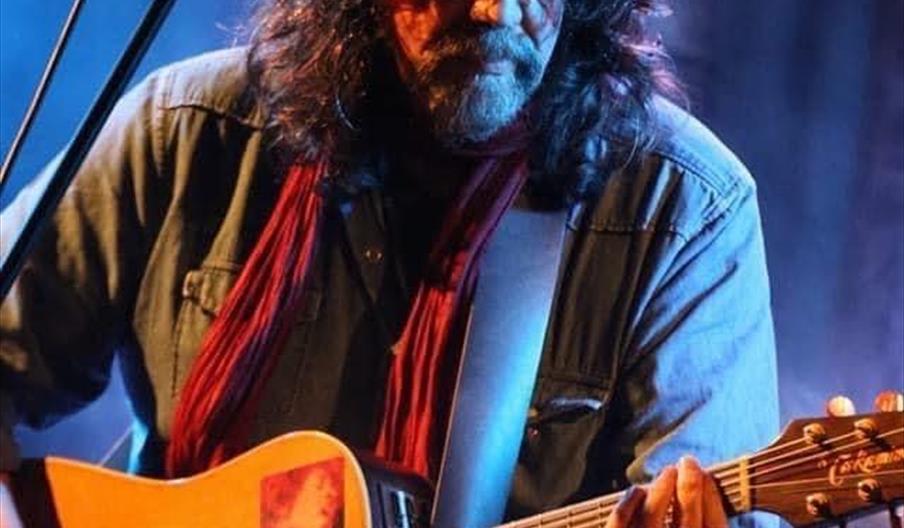 A man with long curly hair and glasses plays an acoustic guitar and sings into a microphone on stage under blue and purple lighting.