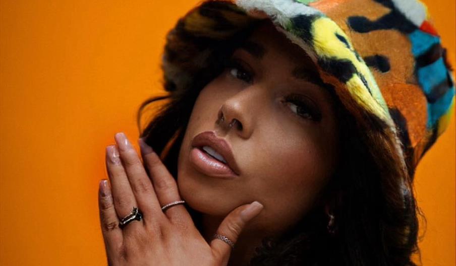 A woman with long dark hair and tattoos on her arms poses against an orange background, wearing a colorful, fuzzy bucket hat and jewelry on her finger