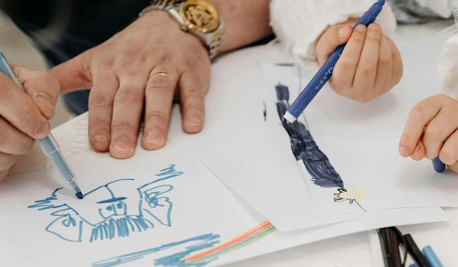 Close-up of an adult and a child drawing together with colored markers on white paper; the adult draws a blue face while the child colors in dark blue