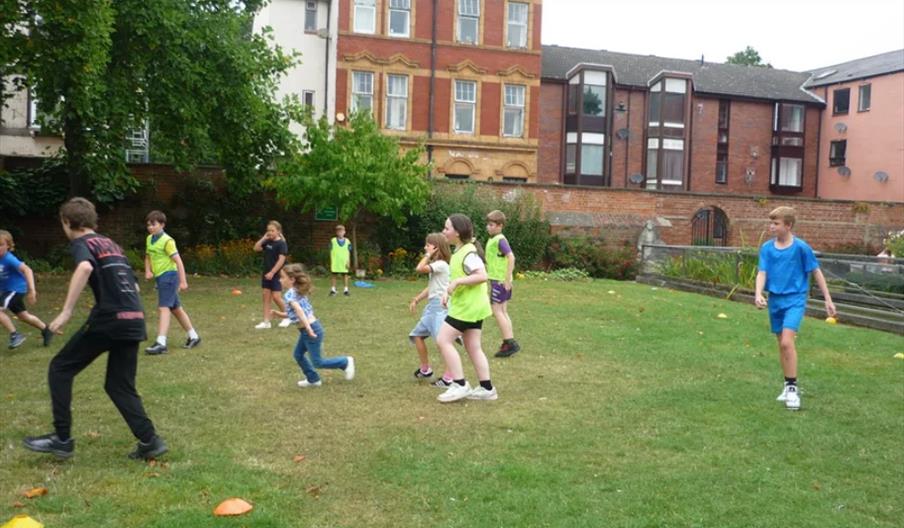 A group of children are playing a game or sport on a grassy field, with some wearing yellow bibs. In the background, there are red-brick and white bui