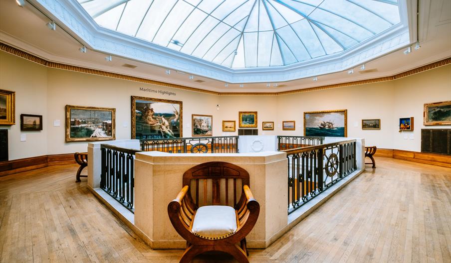 A museum gallery with a large skylight, light wooden floors, maritime-themed paintings on the walls, and ornate wooden benches placed near a railing t