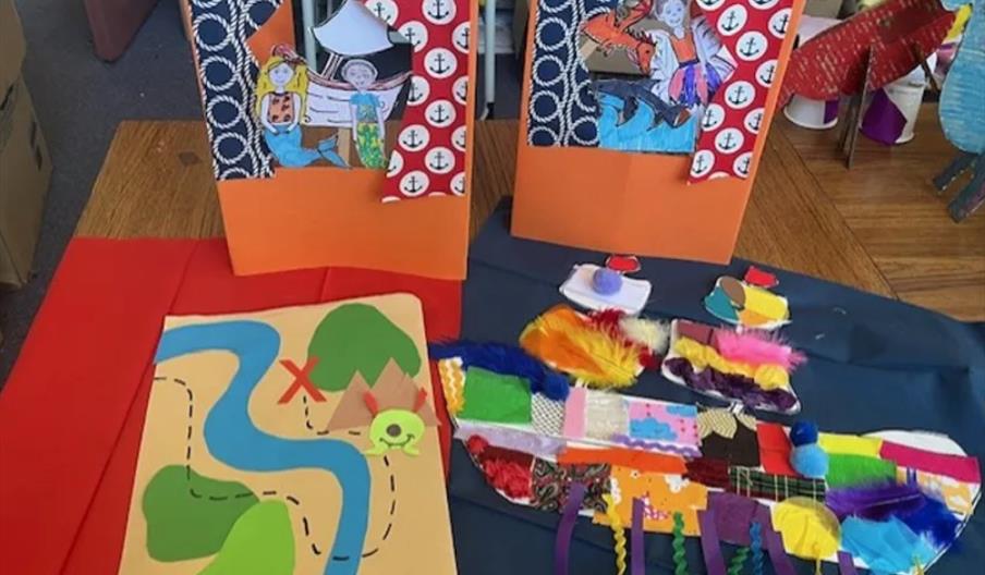 A collection of colorful children's crafts on a table, including a handmade treasure map, a textured collage of a ship using various materials, and tw