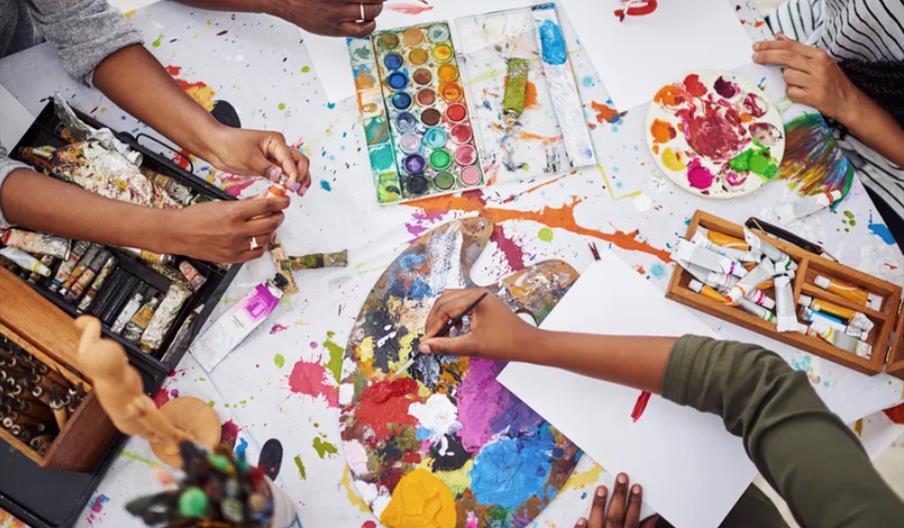 Four people sit around a messy table filled with art supplies, including paint tubes, brushes, watercolor palettes, and paper; three of them are activ