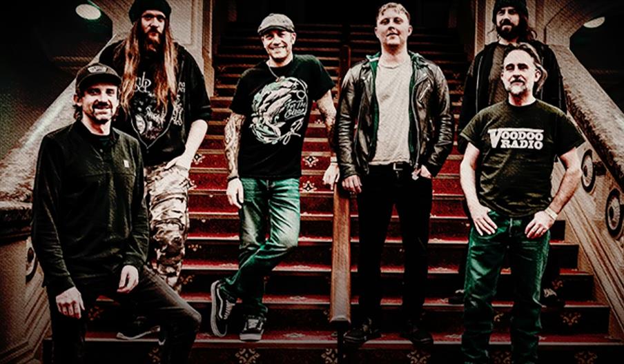 Six men stand and pose on a carpeted staircase in an ornate indoor setting, all wearing casual clothing including t-shirts, jeans, and hats. The group