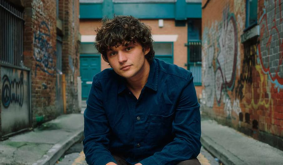 A young man with curly brown hair and a dark blue shirt sits in a narrow alleyway lined with brick walls covered in colorful graffiti. He looks direct