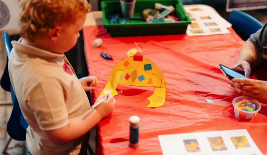 A young child works on a colourful paper crown craft at a table covered with a red tablecloth, while an adult beside them uses a smartphone. Art suppl