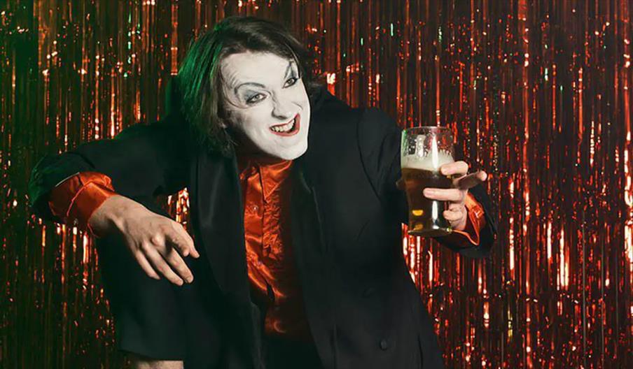 A person with white face paint, dark eye makeup, and a wide grin is wearing a black blazer over a shiny orange shirt, holding a pint of beer and posin