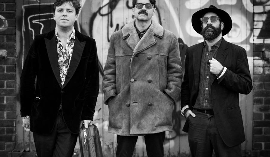 Three men stand in front of a graffiti-covered wall. They are dressed in stylish, vintage-inspired clothing—one wears a velvet blazer and floral shirt