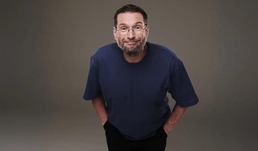 Gary Delaney with glasses, short dark hair, and a beard is standing against a plain gray background, leaning slightly forward with his hands behind hi