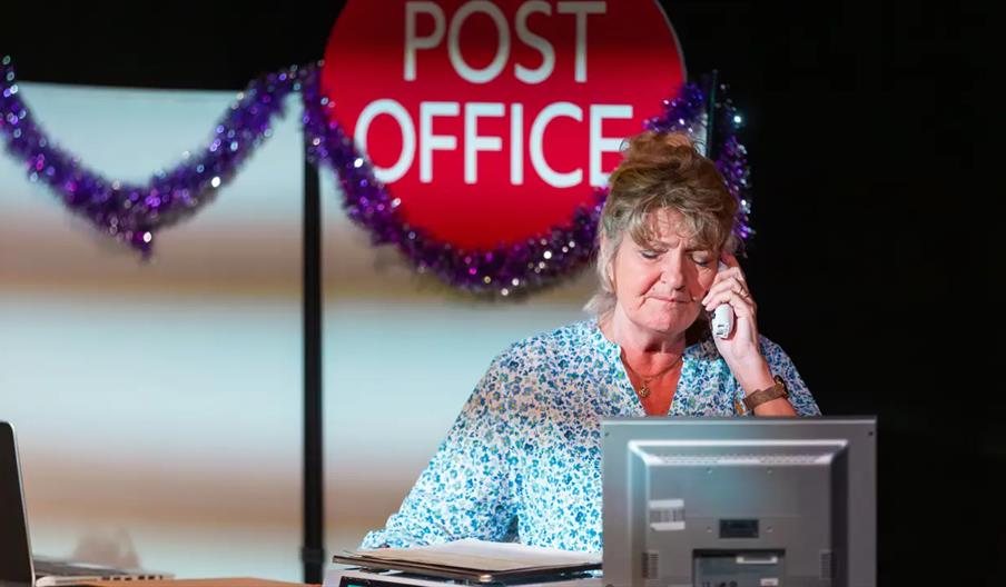 A woman sits at a desk in front of a computer and talks on the phone, with a large "Post Office" sign and purple tinsel decorations in the background.