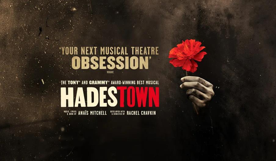 Poster for the musical "Hadestown" featuring the show's title, a hand holding a bright red flower against a dark, textured background, and text descri