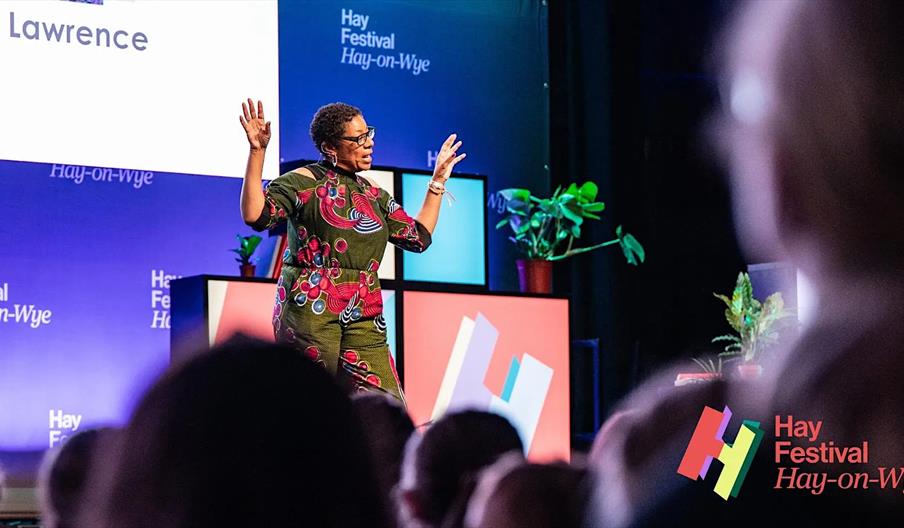 A woman stands on stage speaking to an audience at the Hay Festival Hay-on-Wye, with colorful lighting, background plants, and the festival logo visib
