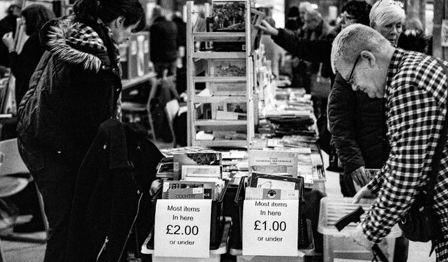 Black and white photo of several people browsing through bins of books at an indoor book sale, with signs indicating prices of £2.00 and £1.00 or unde